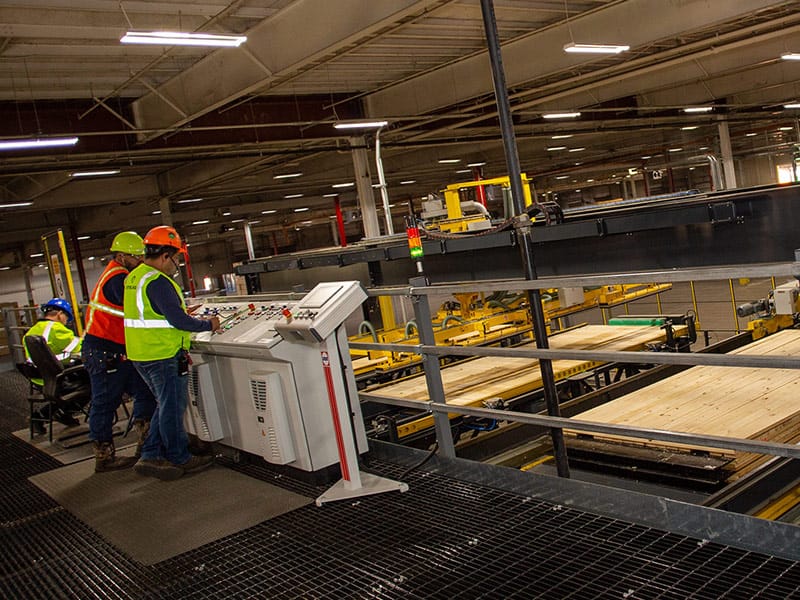 The image shows workers in safety gear operating a control panel at Sterlings Structural Manufacturing facility, with large wooden planks on conveyor systems in the background.