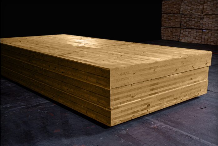 The image shows a large, layered wooden block, made of cross-laminated timber (CLT), in a dark industrial setting with additional timber stacks in the background.