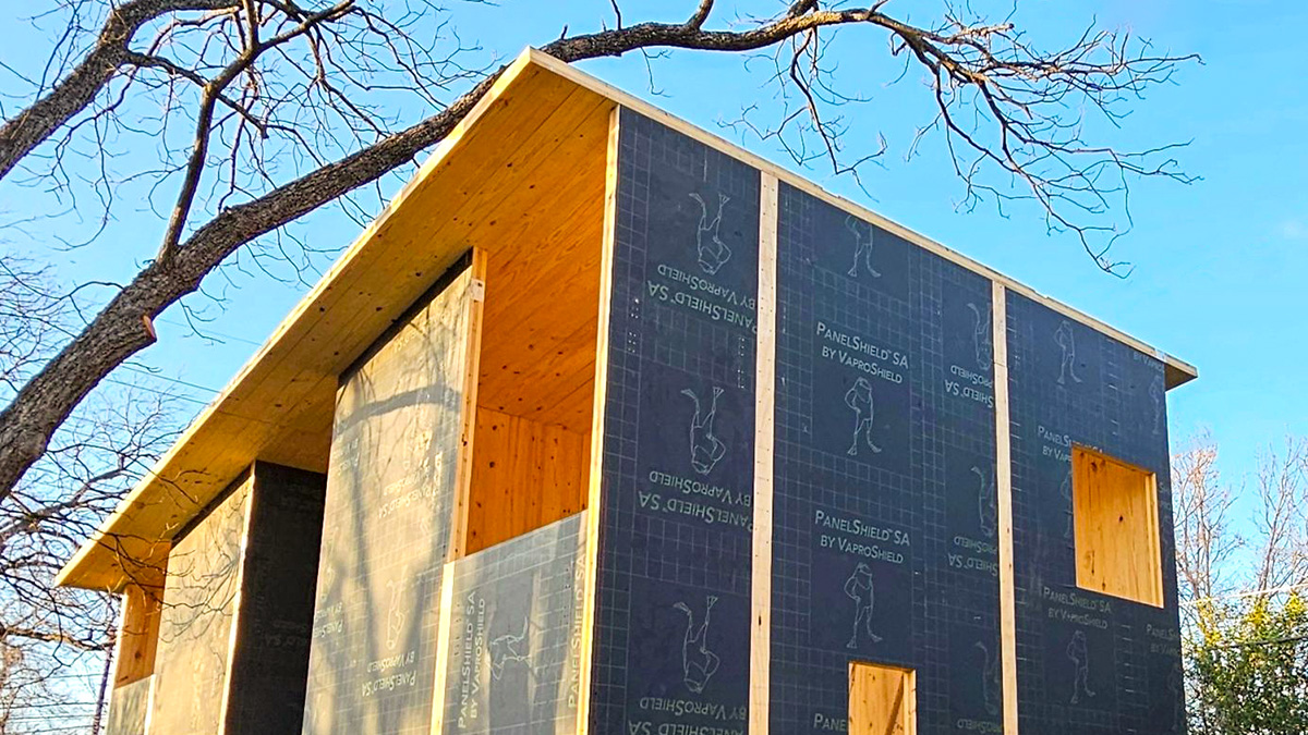 The image shows a partially constructed building with exposed CLT and black sheathing, surrounded by tree branches and overhead wires against a clear blue sky.