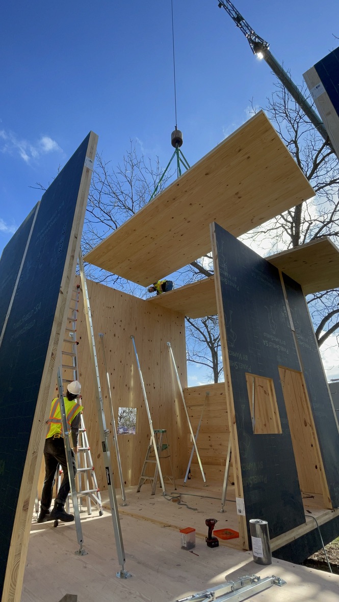 A construction scene showing workers assembling a building using large cross-laminated timber (CLT) panels. A crane is lifting a CLT panel to be placed on top of the structure, while workers in safety gear use ladders and tools to align and secure the panels. The construction is set outdoors on a clear day with a blue sky and trees in the background.