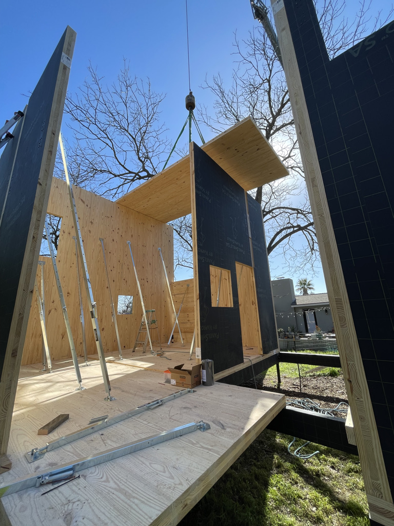 A construction scene showing workers assembling a building using large cross-laminated timber (CLT) panels. A crane is lifting a CLT panel to be placed on top of the structure, while workers in safety gear use ladders and tools to align and secure the panels. The construction is set outdoors on a clear day with a blue sky and trees in the background.