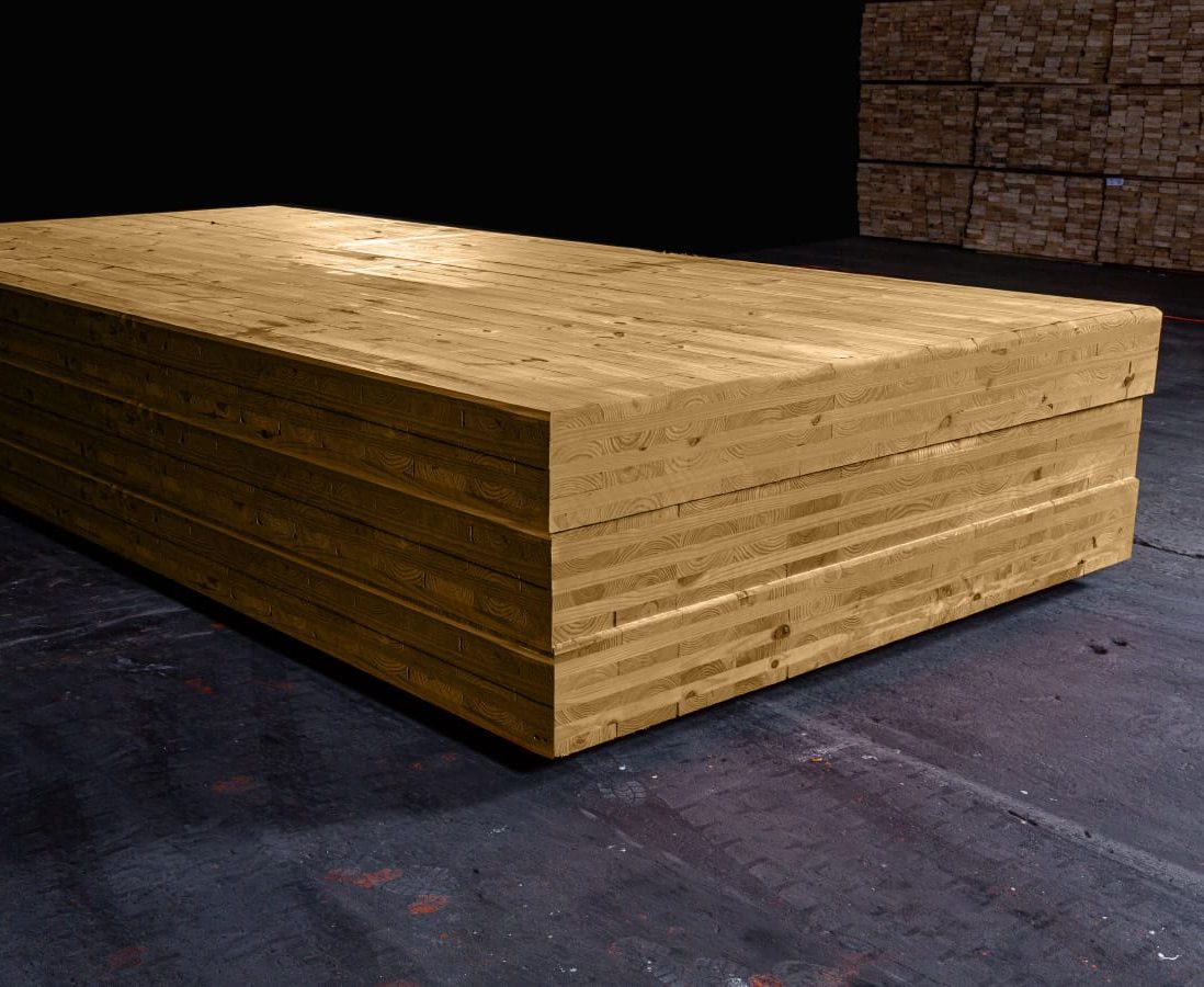 The image shows a large, layered wooden block, made of cross-laminated timber (CLT), in a dark industrial setting with additional timber stacks in the background.