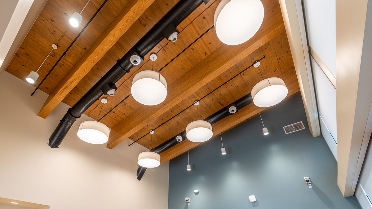 A ceiling with exposed CLT wooden beams and ceiling, featuring round pendant lights and black ductwork. The design combines industrial and natural elements, with the warm wood contrasting against the sleek lighting and duct system.