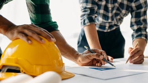 The image shows two people reviewing construction plans with a yellow hard hat and rolled-up blueprints on the table.