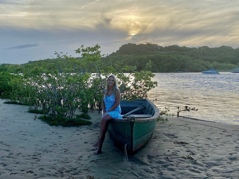 A woman sitting on an old wooden boat on a sandy shore at sunset. She is wearing a blue dress and smiling, with the calm water, mangrove trees, and distant hills creating a serene backdrop.