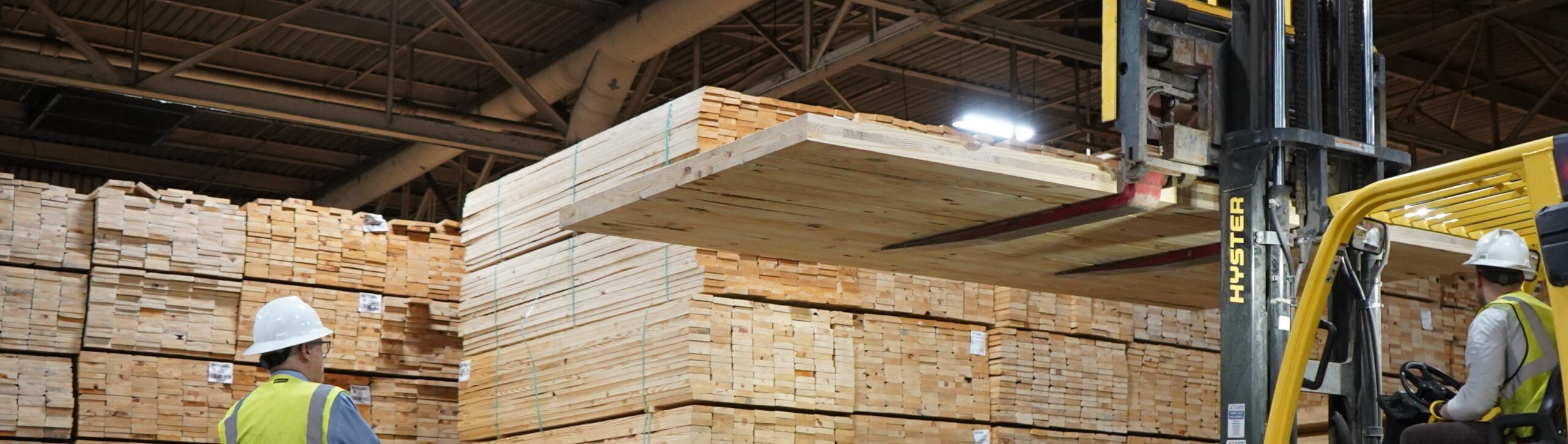 The image shows workers in hard hats and safety vests operating a forklift to move large stacks of lumber in a warehouse. The forklift is lifting a large wooden panel, and numerous stacks of timber are visible in the background.