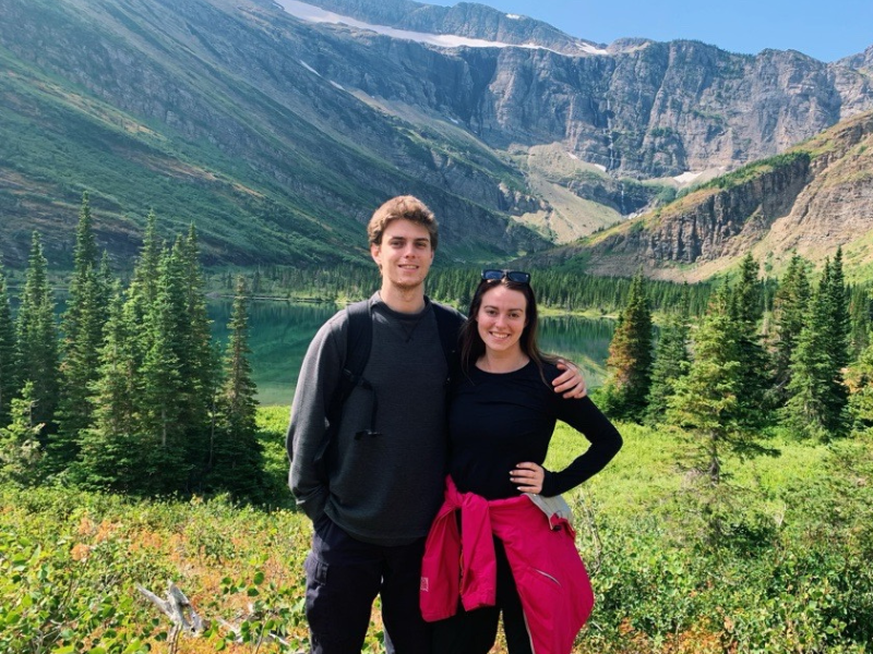 A smiling couple stands together in front of a scenic mountain backdrop with evergreen trees and a calm lake. They are dressed in outdoor clothing, enjoying a clear, sunny day in a lush, natural setting.