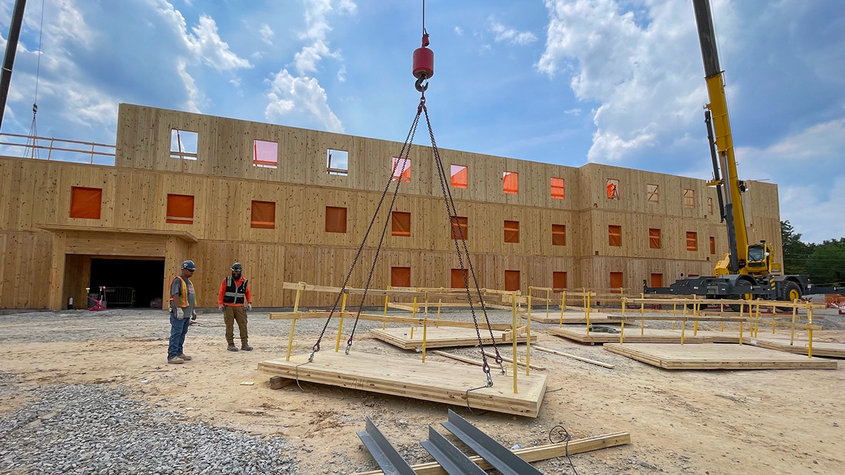 The image shows two workers at a construction site where a crane is lifting a large wooden panel in front of a multi-story wooden building with window openings.
