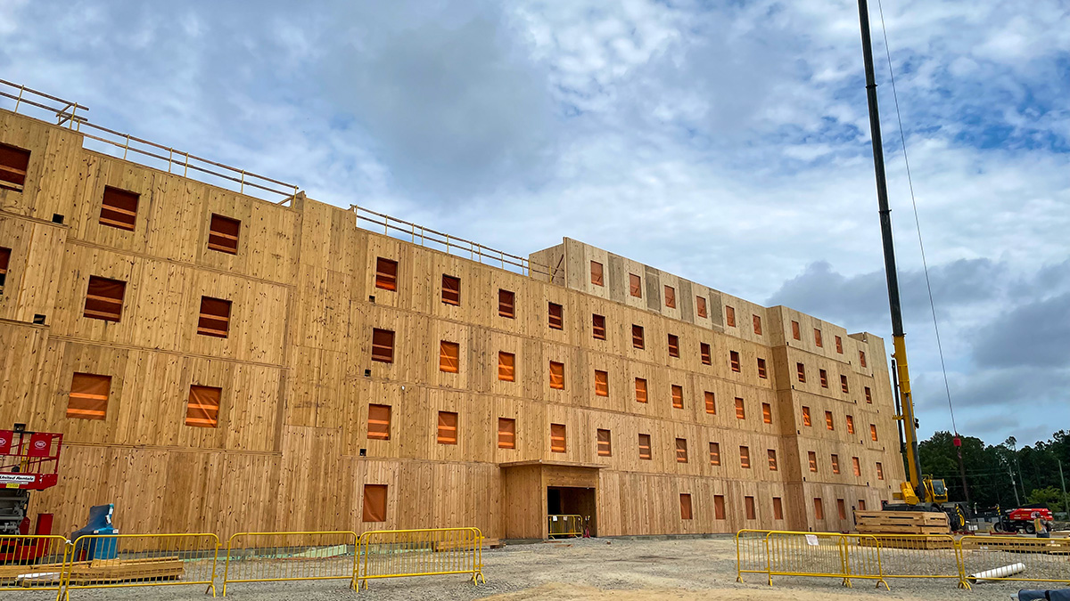 The image shows a large multi-story Mass Timber building under construction, with multiple square window openings. Yellow barriers surround the site, and a crane is visible on the right. The sky is overcast with scattered clouds.