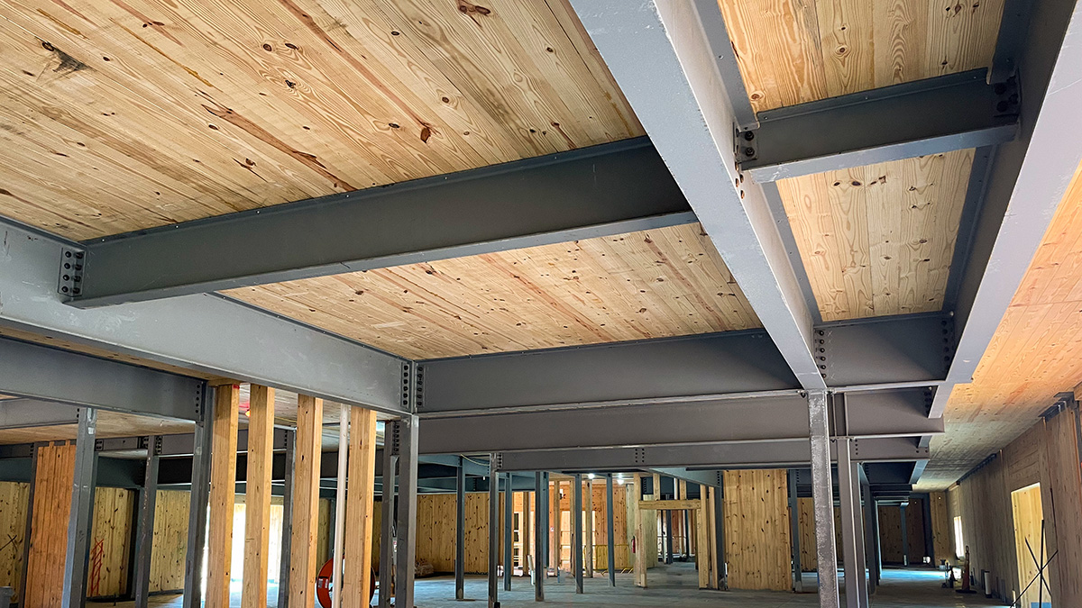 Interior view of a building under construction with CLT ceilings supported by steel beams, showing a hybrid wood and steel structure.
