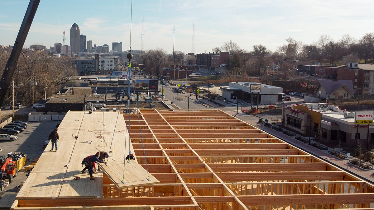 The image shows a construction site with workers installing CLT panels on the upper floor of a building. A crane is lifting materials, and the wooden framework of the structure is clearly visible. The scene is set in an urban area with buildings, roads, and a city skyline in the background.