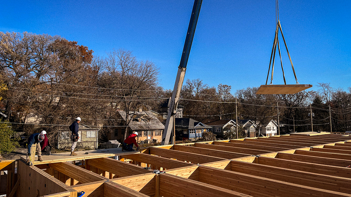 Workers at a construction site guide a crane lifting a mass timber rectangular panel over a building frame, with trees and houses in the background under a clear blue sky.