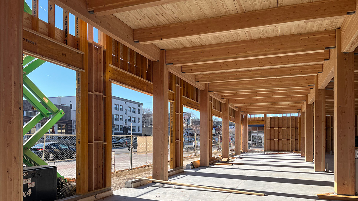 An open timber-framed building under construction, with sunlight casting shadows on the concrete floor through large side openings.