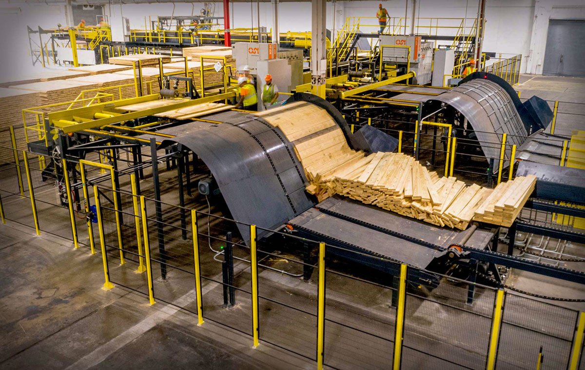 The image shows an Sterlings Lufkin location facility with large curved conveyor belts handling wooden boards. Workers in safety gear oversee the machinery from elevated platforms, and the area is secured with fencing.