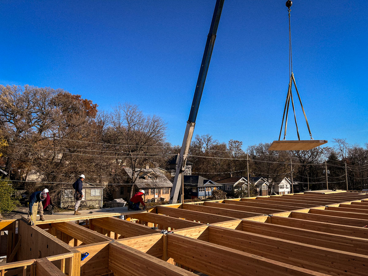 Workers at a construction site guide a crane lifting a mass timber rectangular panel over a building frame, with trees and houses in the background under a clear blue sky.