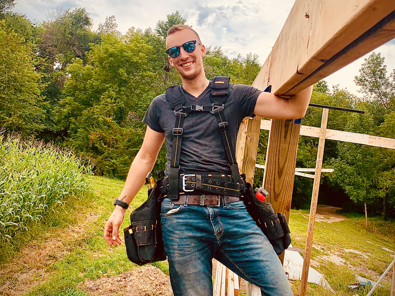 Kyle Teal, wearing a tool belt and sunglasses, smiles while leaning against a wooden structure at a construction site, with trees and greenery in the background.