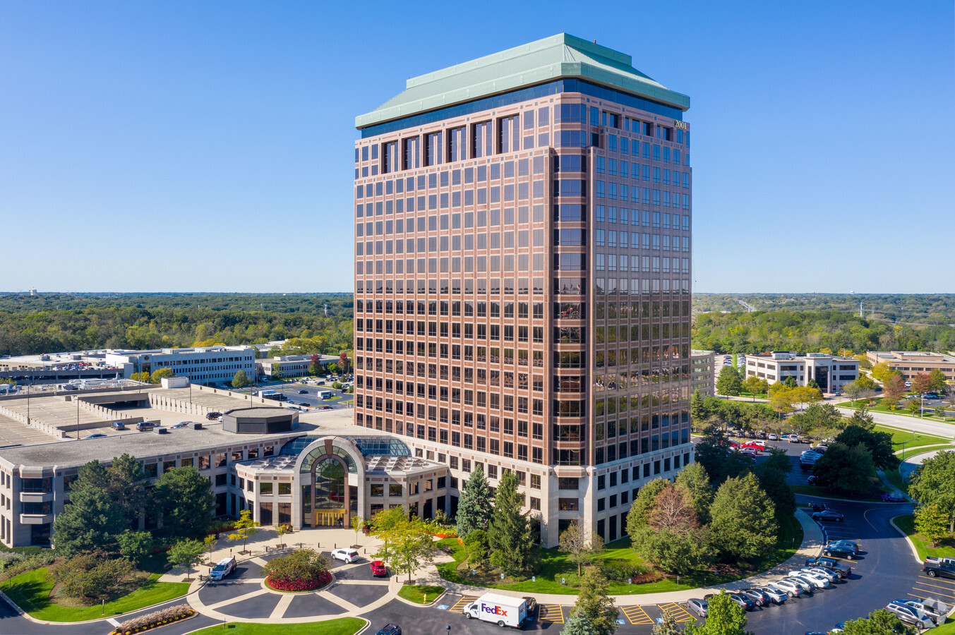 The image shows a tall office building with a glass exterior, arched entrance, and surrounding trees and parking lot, under a clear blue sky.