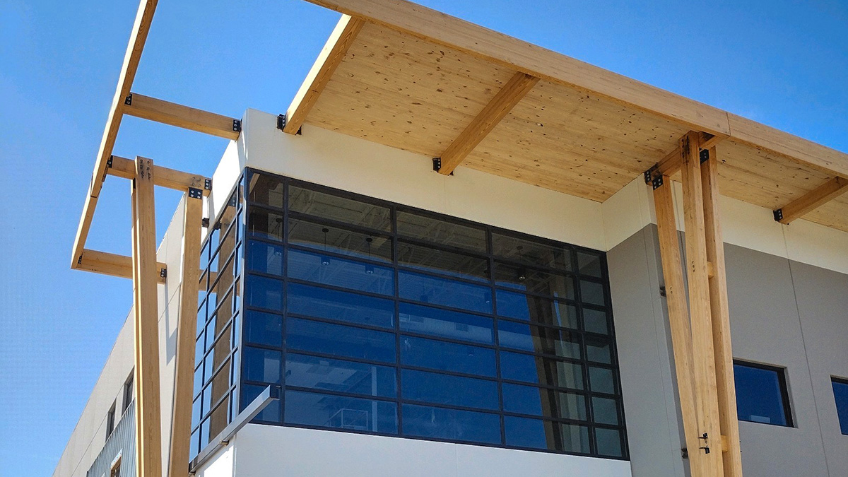 A modern building facade with exposed wooden beams and a CLT roof, featuring glass windows and gray panels.