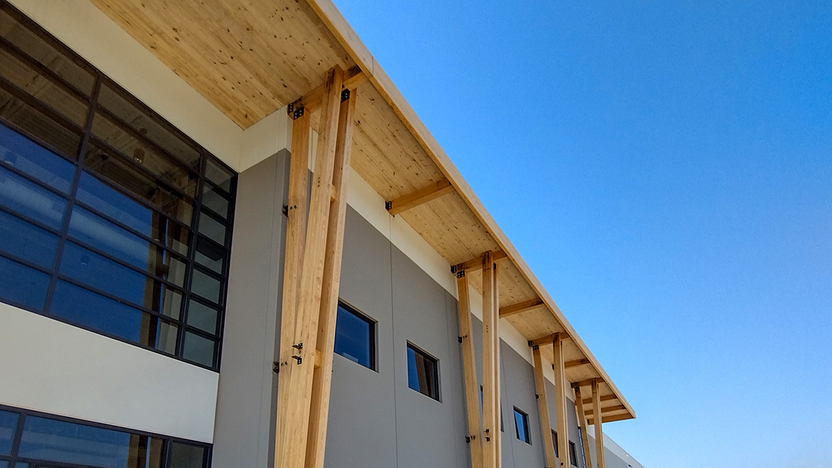 A modern building facade with exposed wooden beams and a CLT roof, featuring glass windows and gray panels.