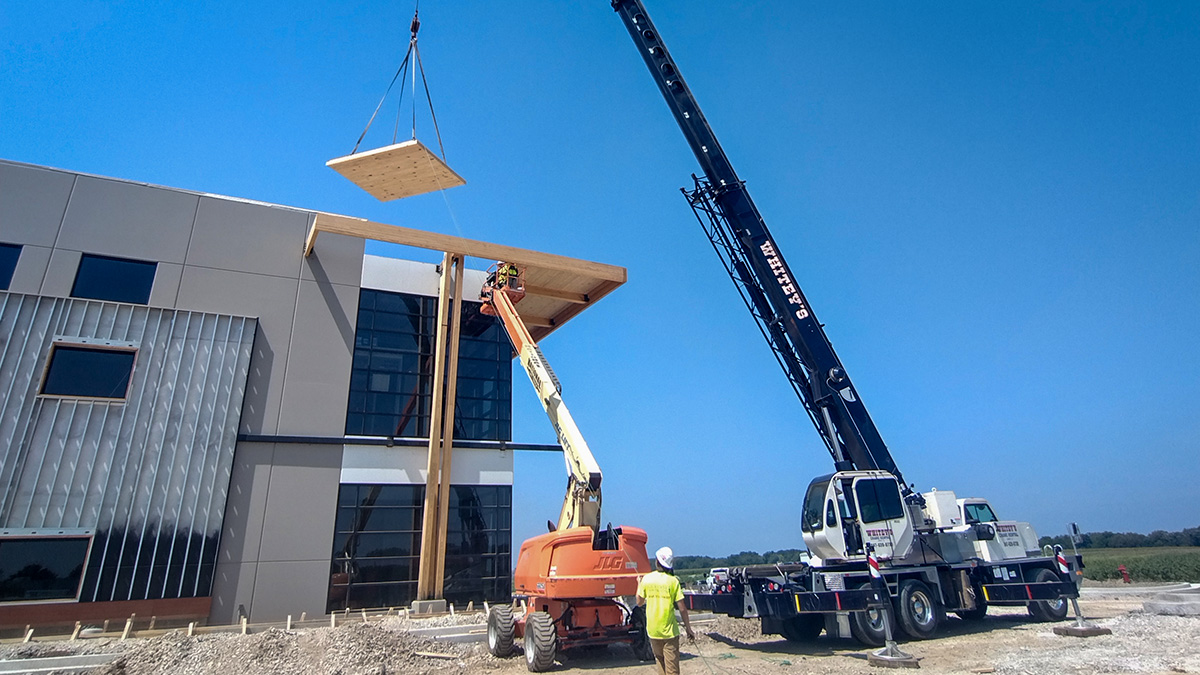 A crane lifts a CLT panel onto a modern building's roof as workers oversee the installation.