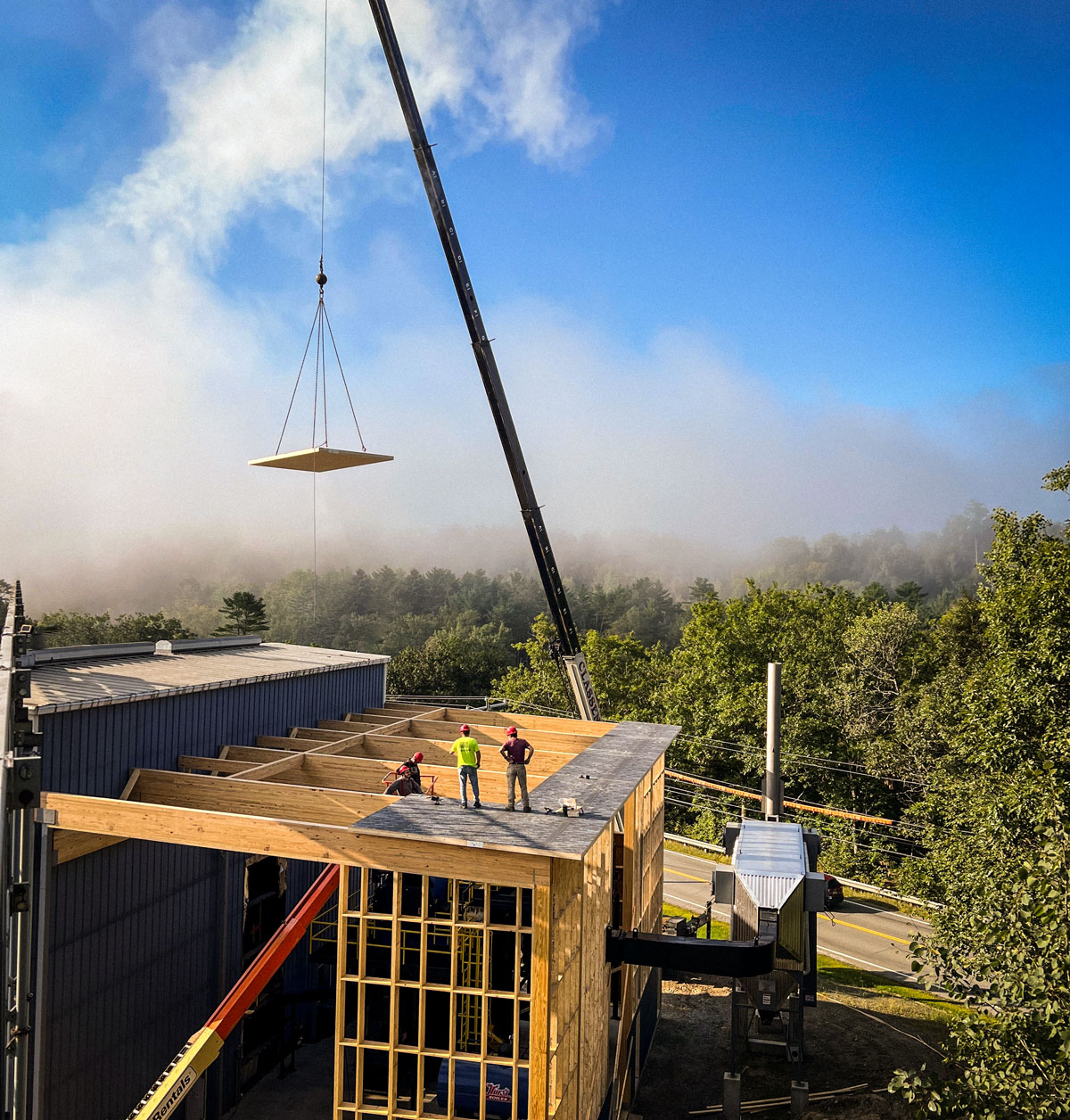 Three workers on a building roof as a crane lifts a mass timber CLT panel, with beautiful trees and mist in the background.