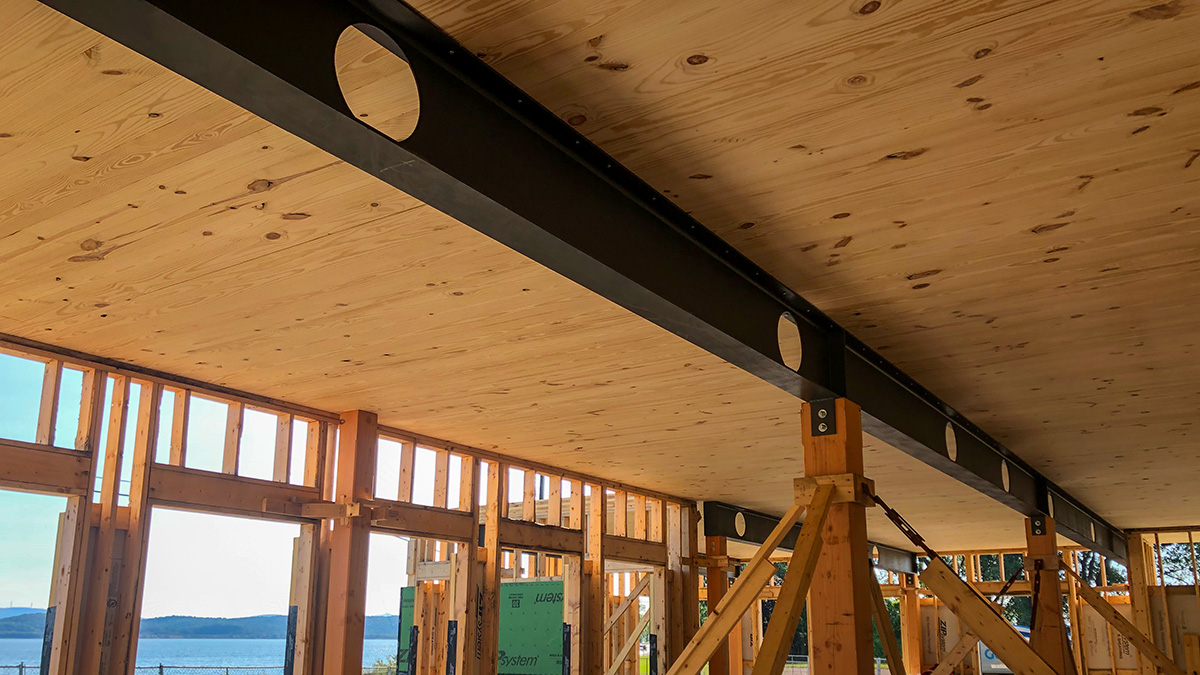 Interior view of a building under construction with CLT ceilings and steel beams with circular cutouts, overlooking water and hills.