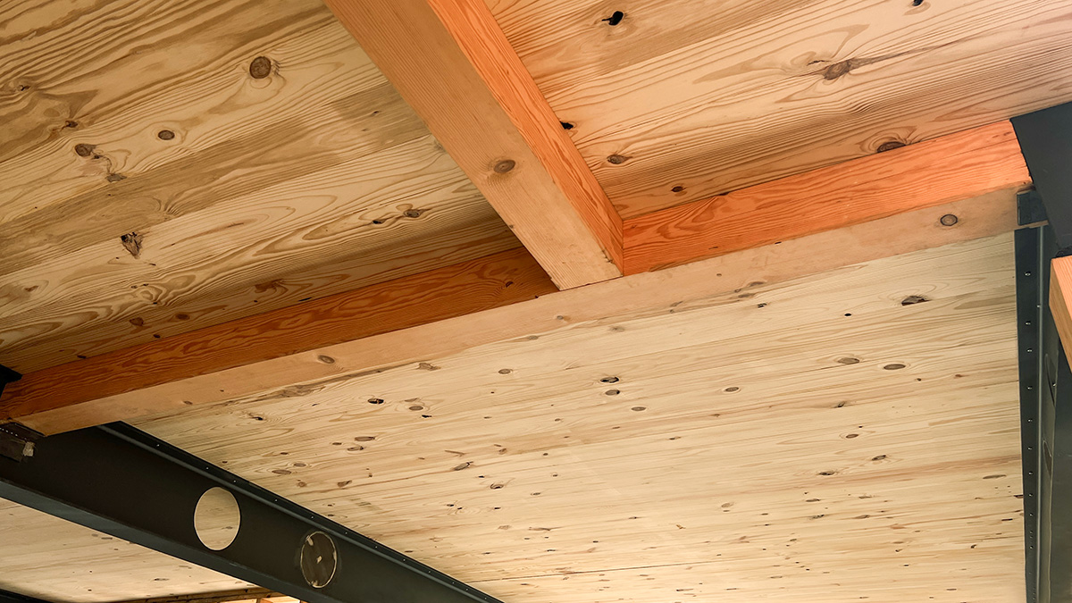 A close-up view of a ceiling with cross-laminated timber (CLT) panels supported by exposed wooden beams and steel framing. The natural wood grain is visible, showcasing knots and textures, while the steel beam features circular cutouts for utilities.