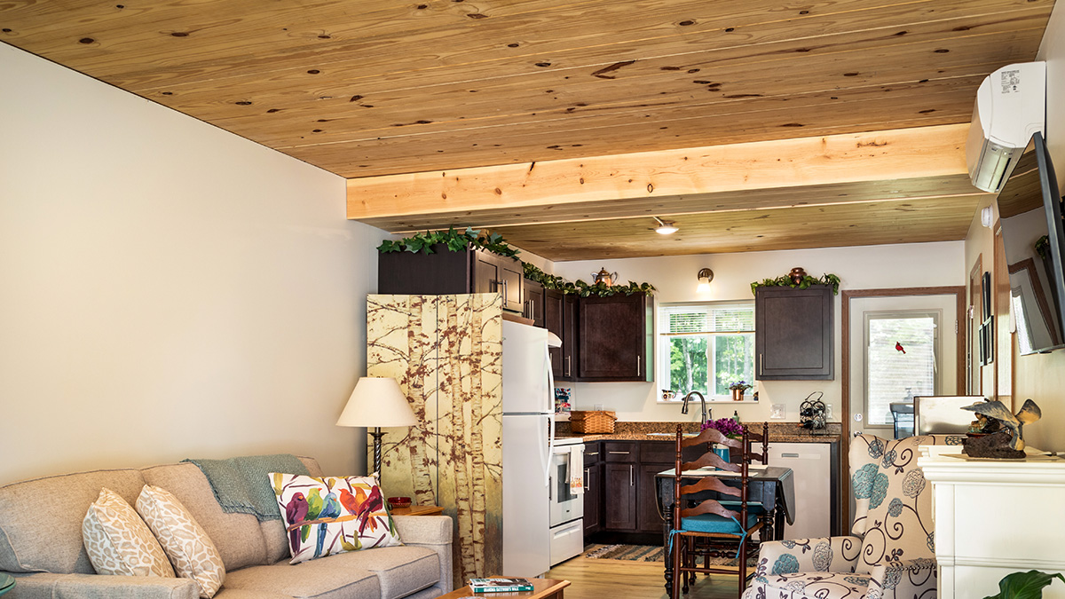 The image shows a cozy living and kitchen area with a wooden ceiling, a couch, colorful pillows, and a dining table. The kitchen has dark cabinets and white appliances.