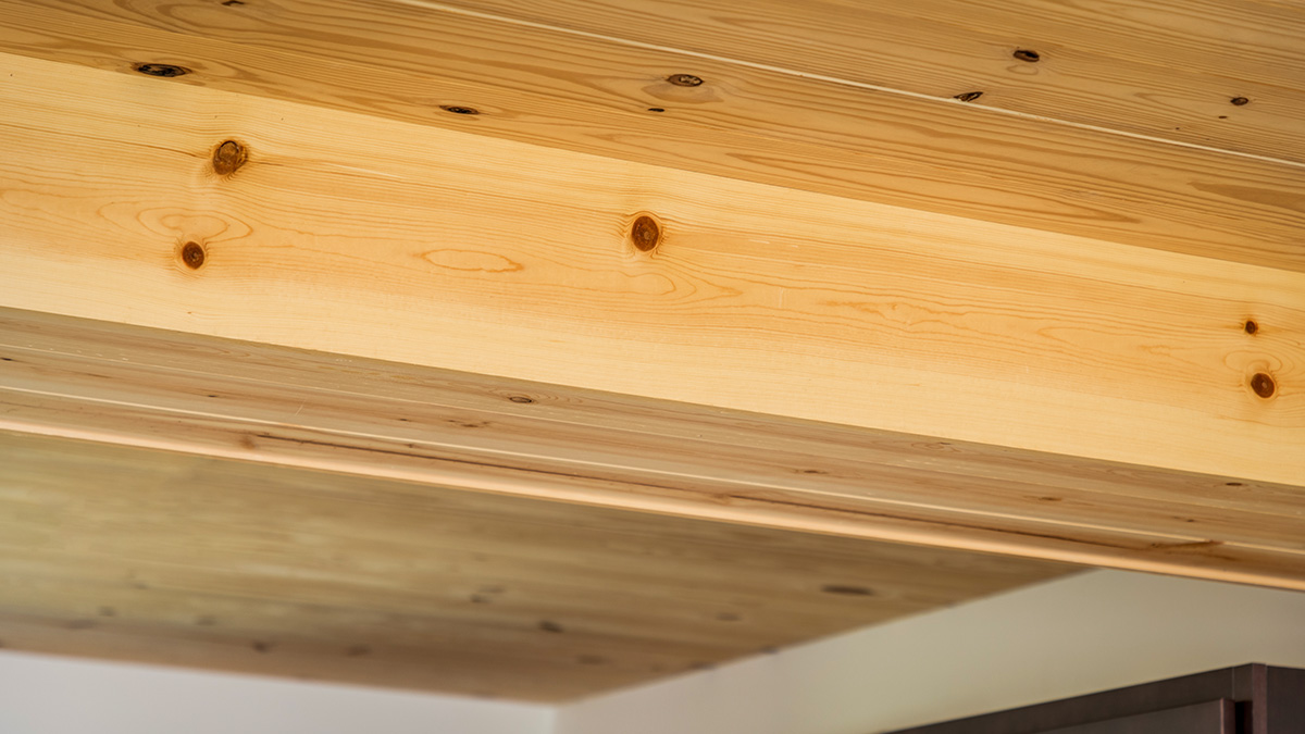 A close-up of a cross laminated timber (CLT) ceiling with visible natural knots and wood grain, showcasing the texture and warm tones of the wood. The ceiling beams create a structured, minimalist design.