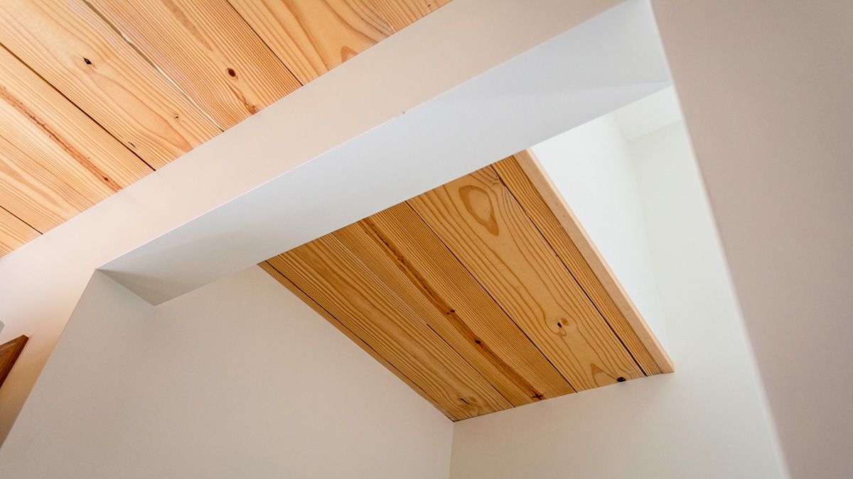 The image shows an interior ceiling detail featuring exposed cross-laminated timber (CLT) set within a white, minimalistic frame. The natural wood grain contrasts with the smooth, white walls, creating a clean, modern look.