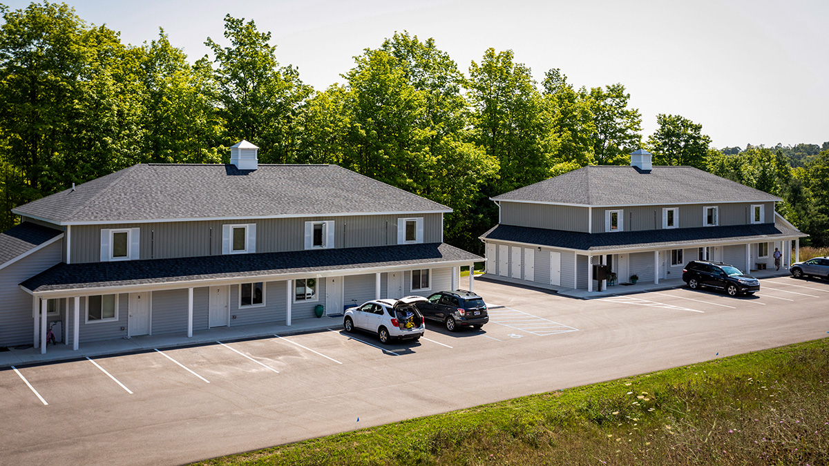 Two identical buildings with gray siding and black roofs sit side by side in a parking lot, each with a few parked cars. The buildings are surrounded by greenery and trees under a clear sky, creating a peaceful suburban setting.