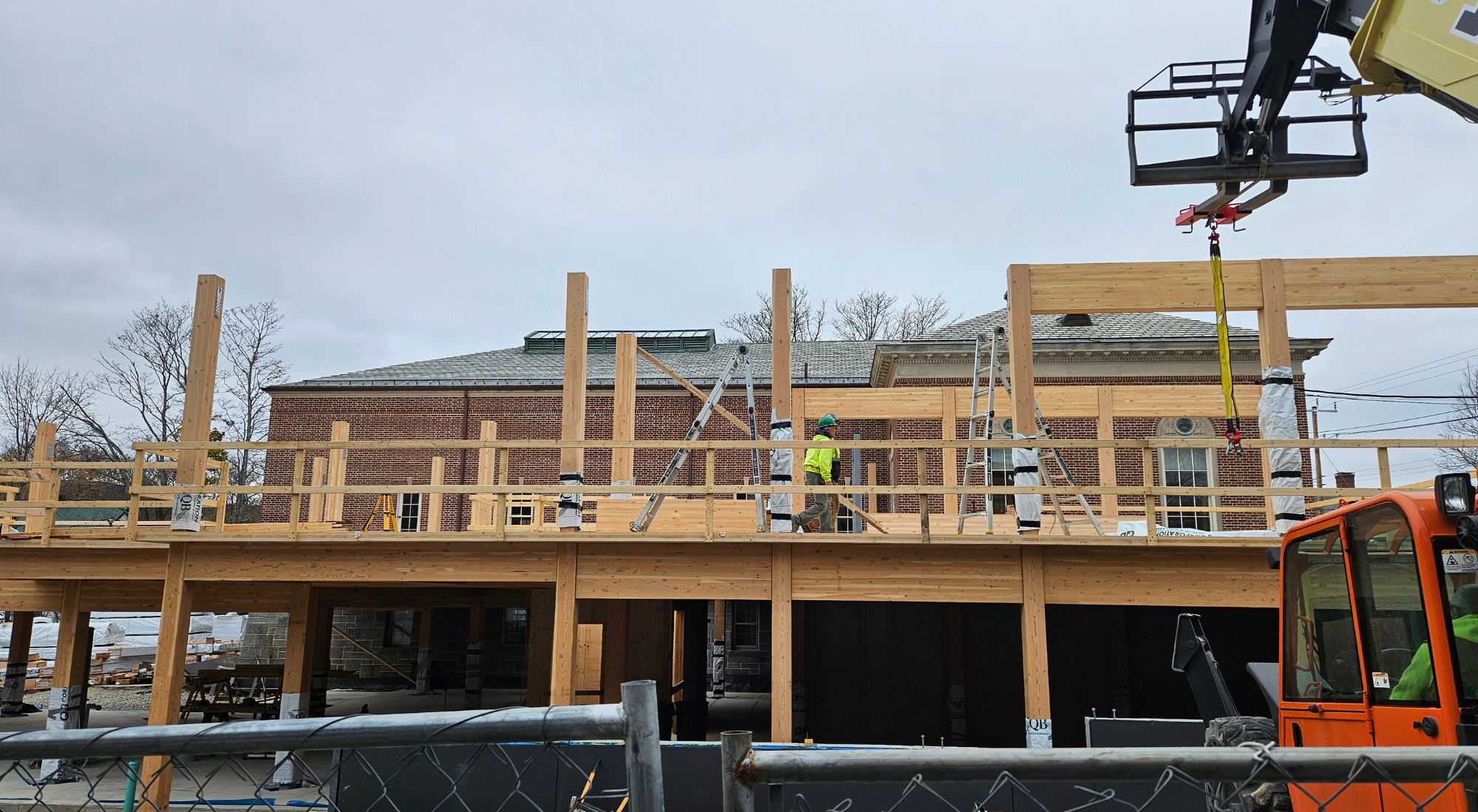 A construction site shows workers assembling a cross-laminated timber (CLT) and glulam structure for an addition to a historic brick library building. The framework includes wooden beams, columns, and scaffolding, with a crane in operation and construction materials visible in the foreground. The brick library stands in the background, surrounded by leafless trees, under a cloudy sky.