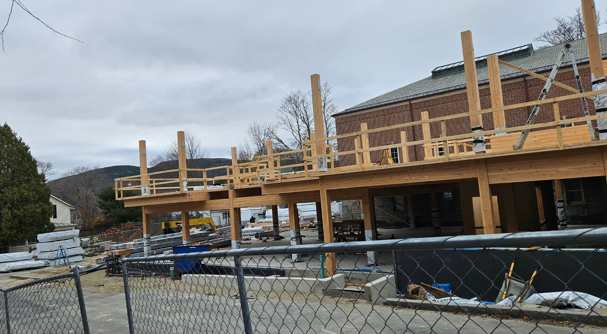 A construction site shows workers assembling a cross-laminated timber (CLT) and glulam structure for an addition to a historic brick library building. The framework includes wooden beams, columns, and scaffolding, with a crane in operation and construction materials visible in the foreground. The brick library stands in the background, surrounded by leafless trees, under a cloudy sky.