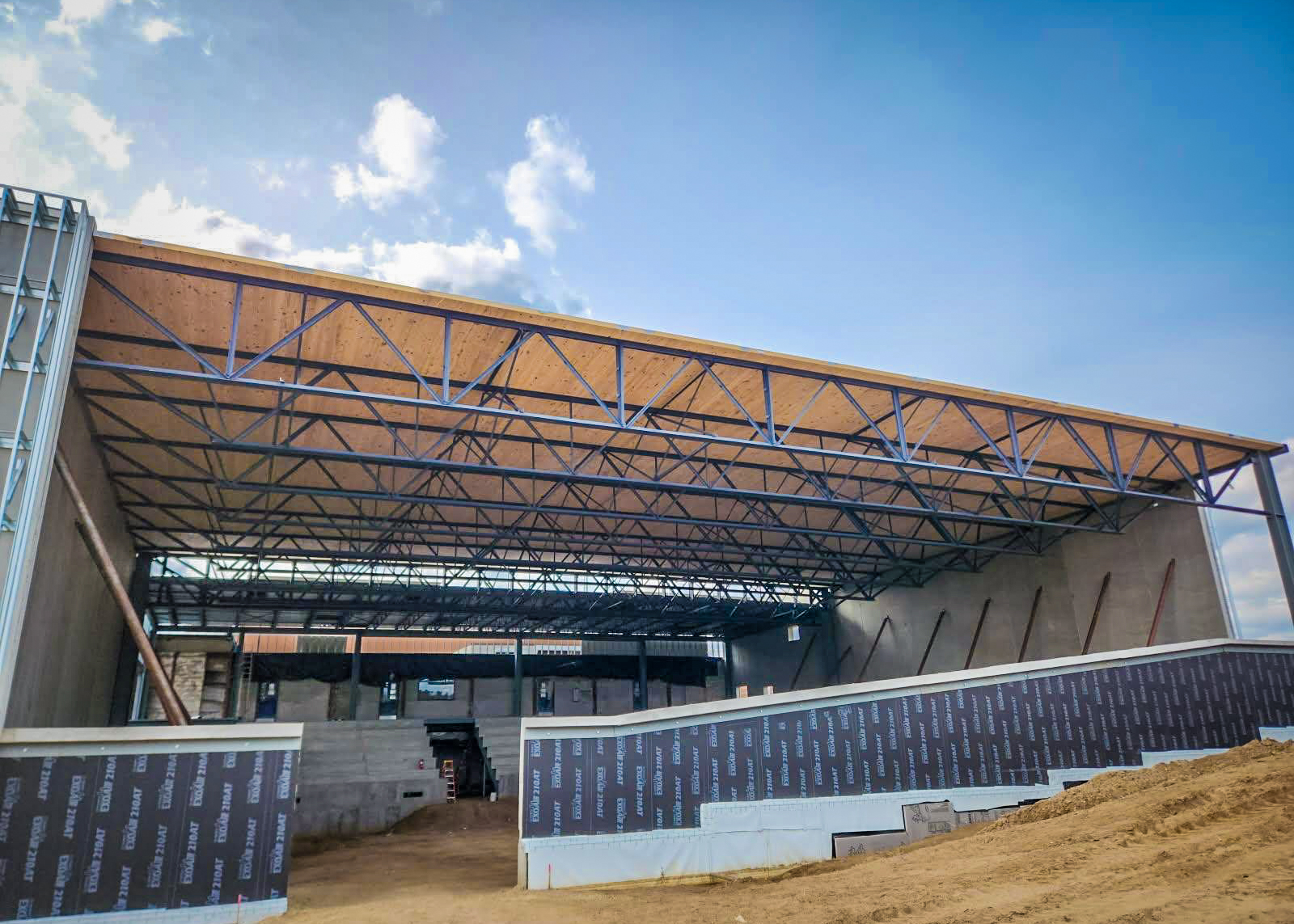 A partially completed natatorium structure at Saint Thomas Academy in Mendota Heights, MN, featuring a steel truss system with cross-laminated timber (CLT) decking being installed. The open framework reveals the construction progress under a bright blue sky.