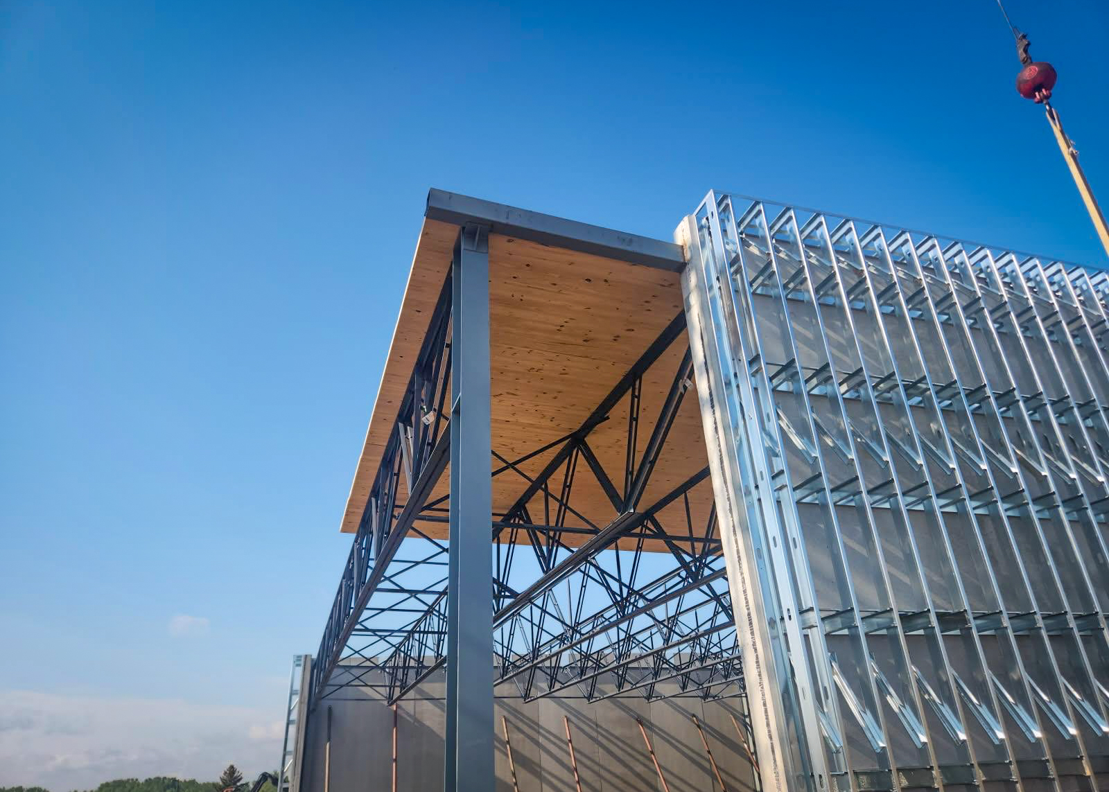 A partially completed natatorium structure at Saint Thomas Academy in Mendota Heights, MN, featuring a steel truss system with cross-laminated timber (CLT) decking being installed. The open framework reveals the construction progress under a bright blue sky.